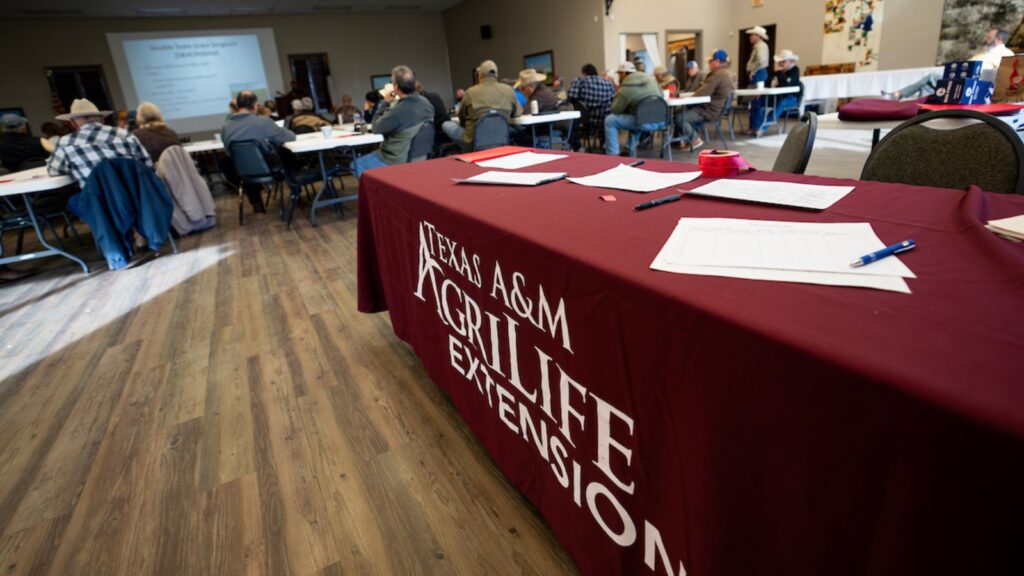 A maroon Texas A&M AgriLife Extension Service registration table with a crowd of people sitting at long tables viewing a slide presentation out of focus in the background